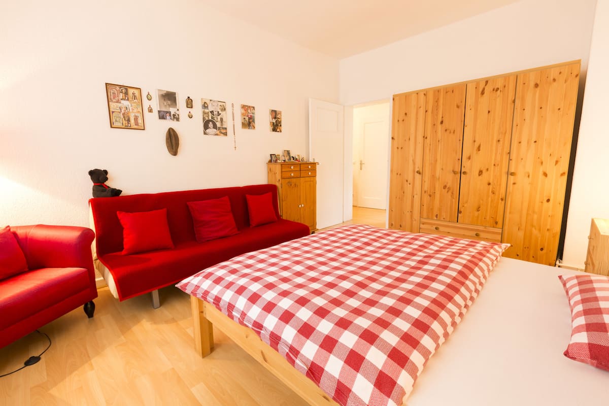 A room with a red sofa, a wooden wardrobe, a checkered bedspread, and a wall with framed pictures.