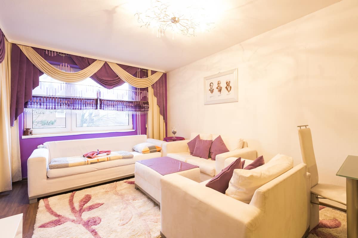 A living room with a sofa set, a coffee table, and a side table. There is a window with curtains, a chandelier, and a framed picture on the wall.