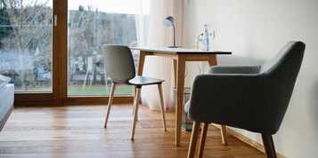 Una habitación con piso de madera, un escritorio con lámpara, dos sillas y una ventana con vista a árboles y casas.