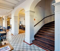 Un vestíbulo de hotel con suelo de cuadros, escalera de madera y un comedor al fondo.