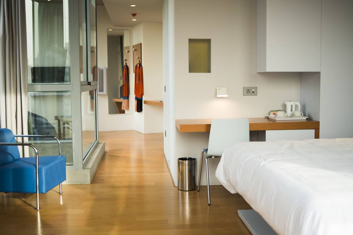 Quarto de hotel com uma cama, uma escrivaninha, uma cadeira e um cesto de lixo.