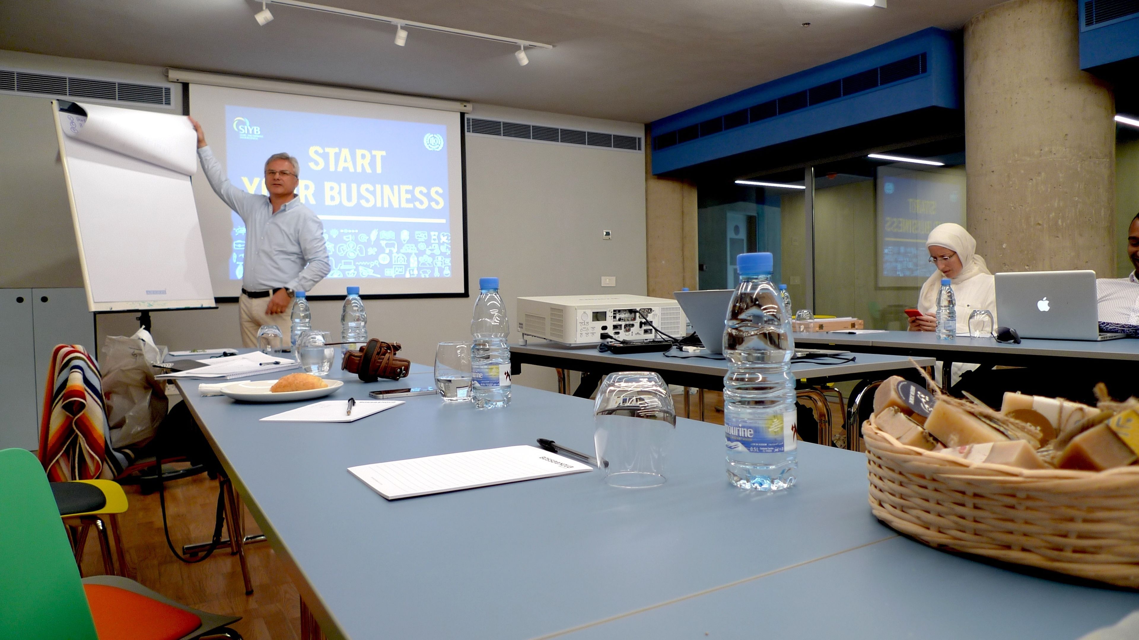 A conference room with a presentation screen displaying "START BUSINESS", a projector, laptops, water bottles, and a basket of bread.