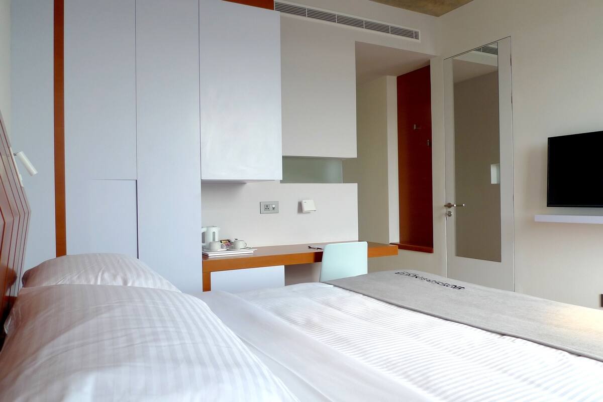 Quarto de hotel com cama, escrivaninha com cadeira, televisão e guarda-roupa.