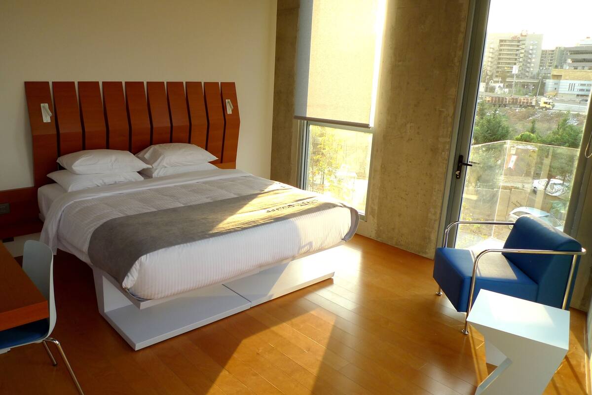 Quarto de hotel moderno com uma cama grande, uma mesa de trabalho, uma cadeira e vista para a cidade através de uma janela ampla.