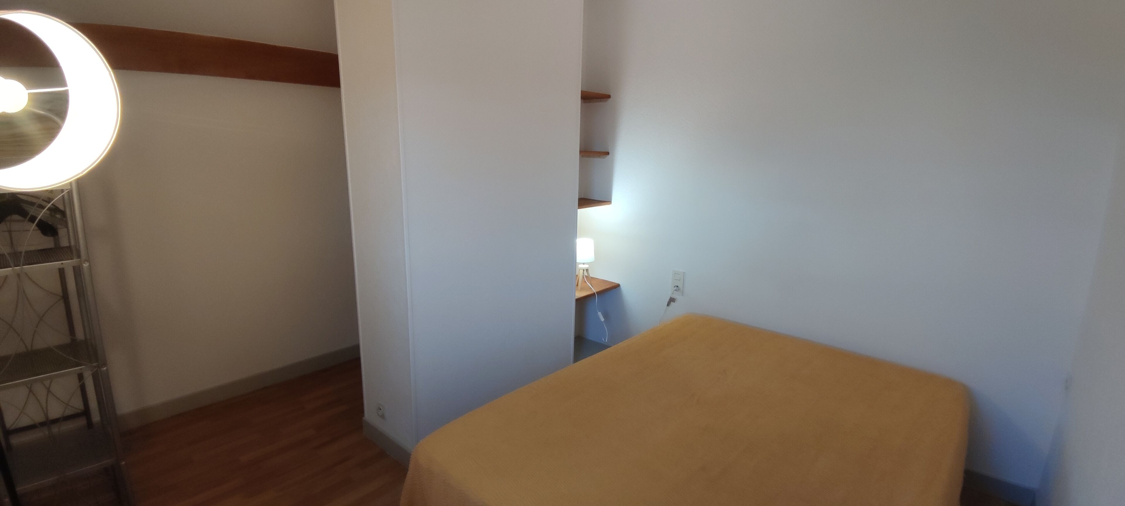 A room with a bed, a standing lamp, a shelf, and a wall socket.