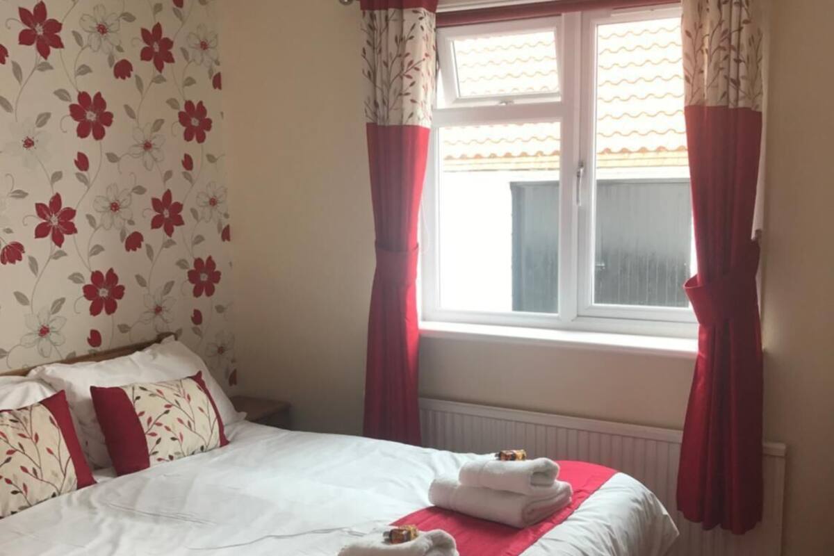 Una habitació d'hotel amb un llit, cortines vermelles, una finestra i paper pintat amb motius florals.