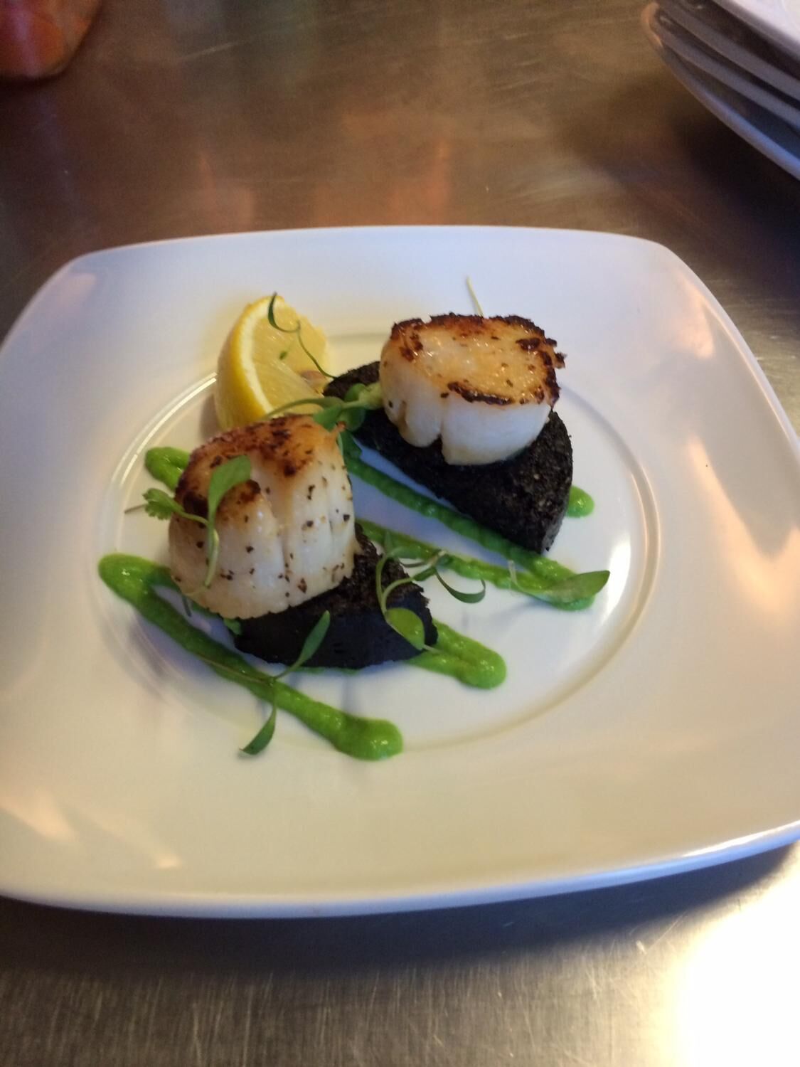 A white plate with two seared scallops on a bed of greens, accompanied by a lemon wedge.