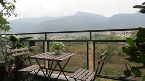 Un balcón con mesa y sillas de madera, plantas en macetas y vistas a montañas y edificios a lo lejos.