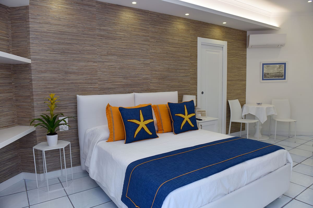 Een moderne hotelkamer met een groot bed, een klein nachtkastje, een pot met een plant en een zithoek met een tafel en stoelen.