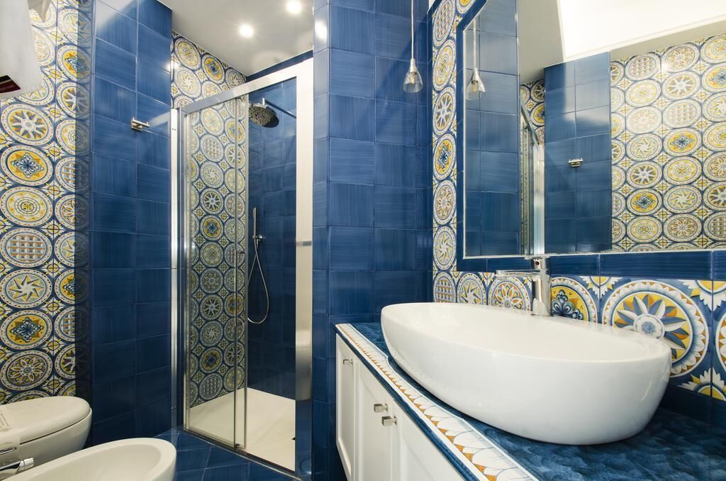 Un baño con un lavamanos grande de color blanco, una cabina de ducha con mampara de vidrio y baldosas azules con diseño.