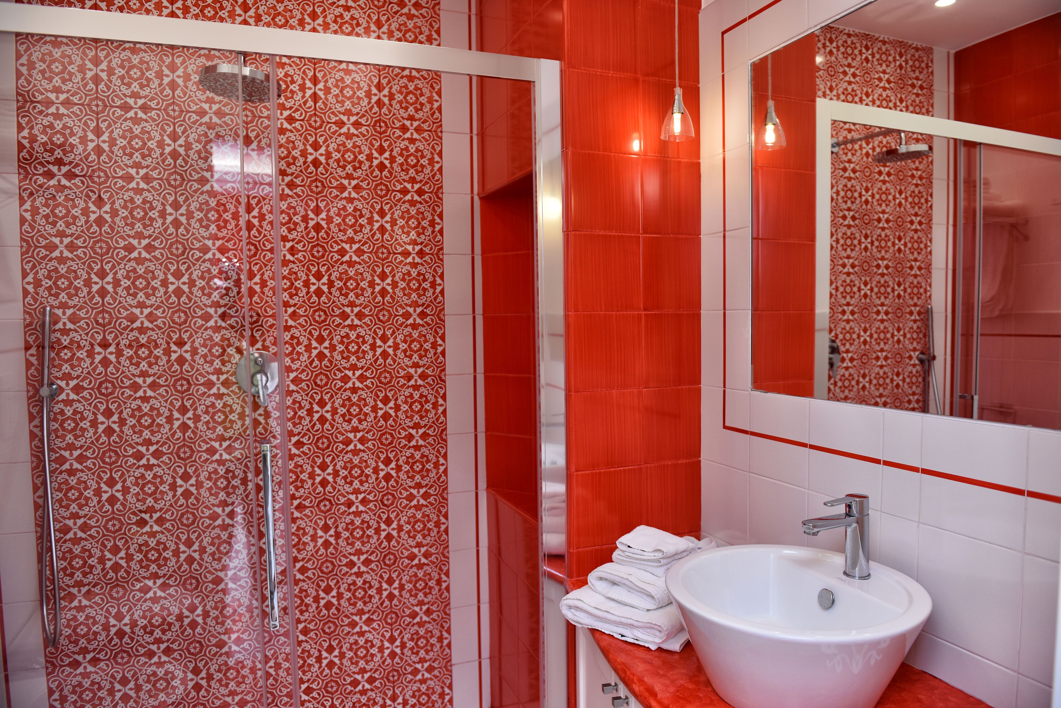 Une salle de bain avec une douche à parois de verre, un lavabo blanc et un miroir qui reflète les carreaux rouges à motifs.