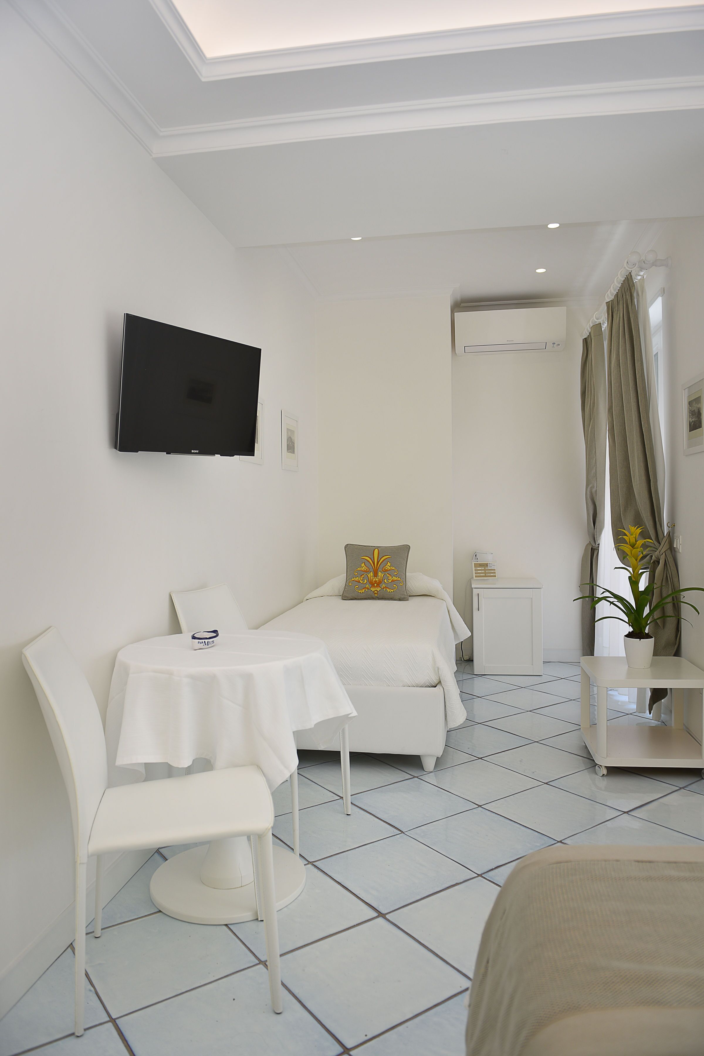 Una habitación de hotel con cama, una mesita, una silla, un televisor montado en la pared, una planta en maceta y una ventana con cortinas.