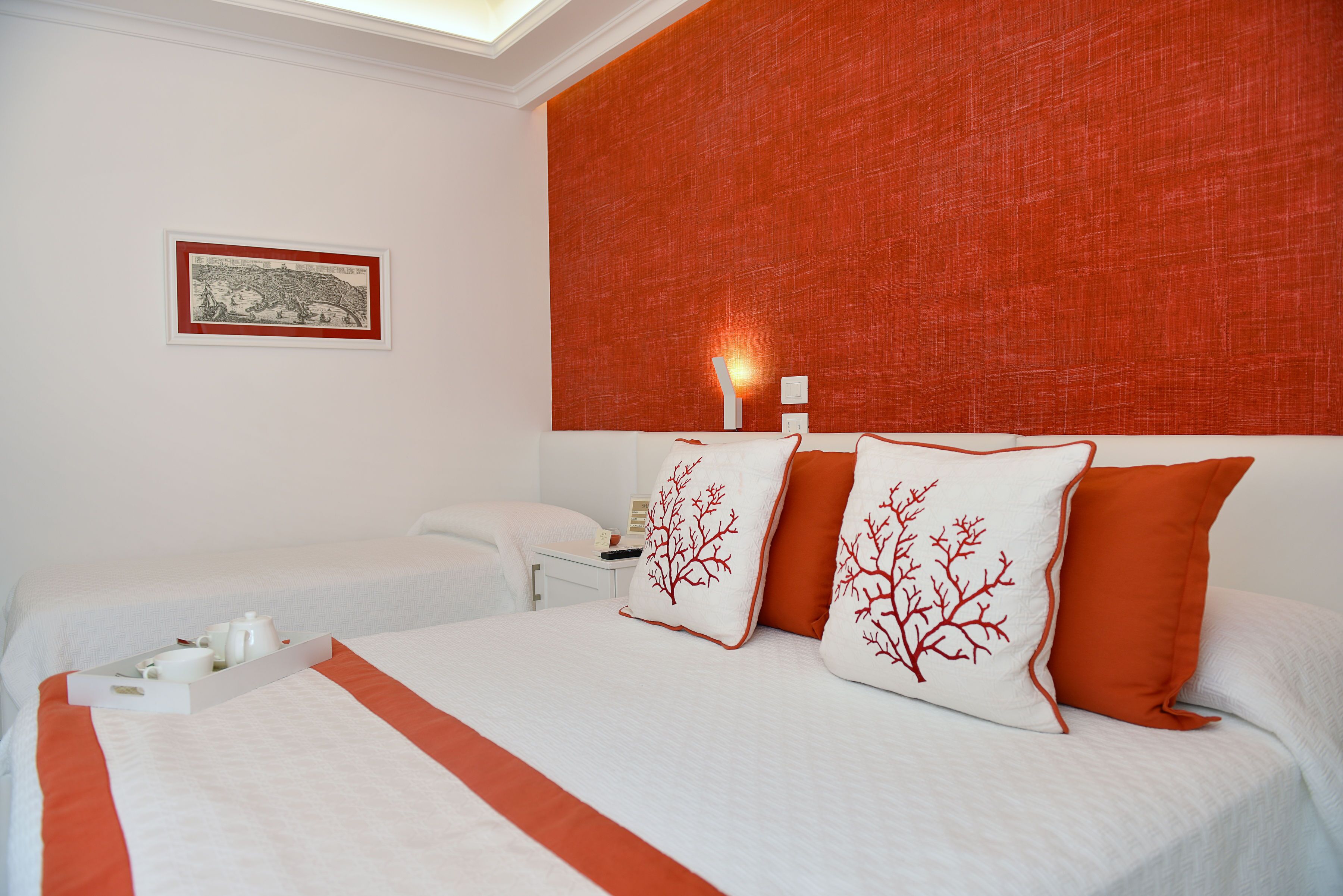 Une chambre d’hôtel avec un grand lit, deux oreillers ornés de motifs d’arbres rouges, une table de chevet avec une lampe et un tableau encadré au mur.