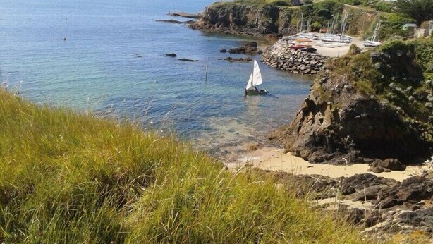 Un paysage côtier avec un voilier sur l’eau, des falaises rocheuses et un petit bâtiment sur la plage.