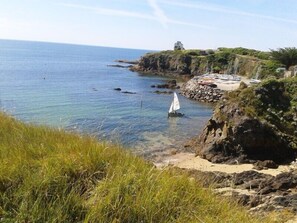 Un paysage côtier avec un voilier sur l’eau, des falaises rocheuses et un petit bâtiment sur la plage.