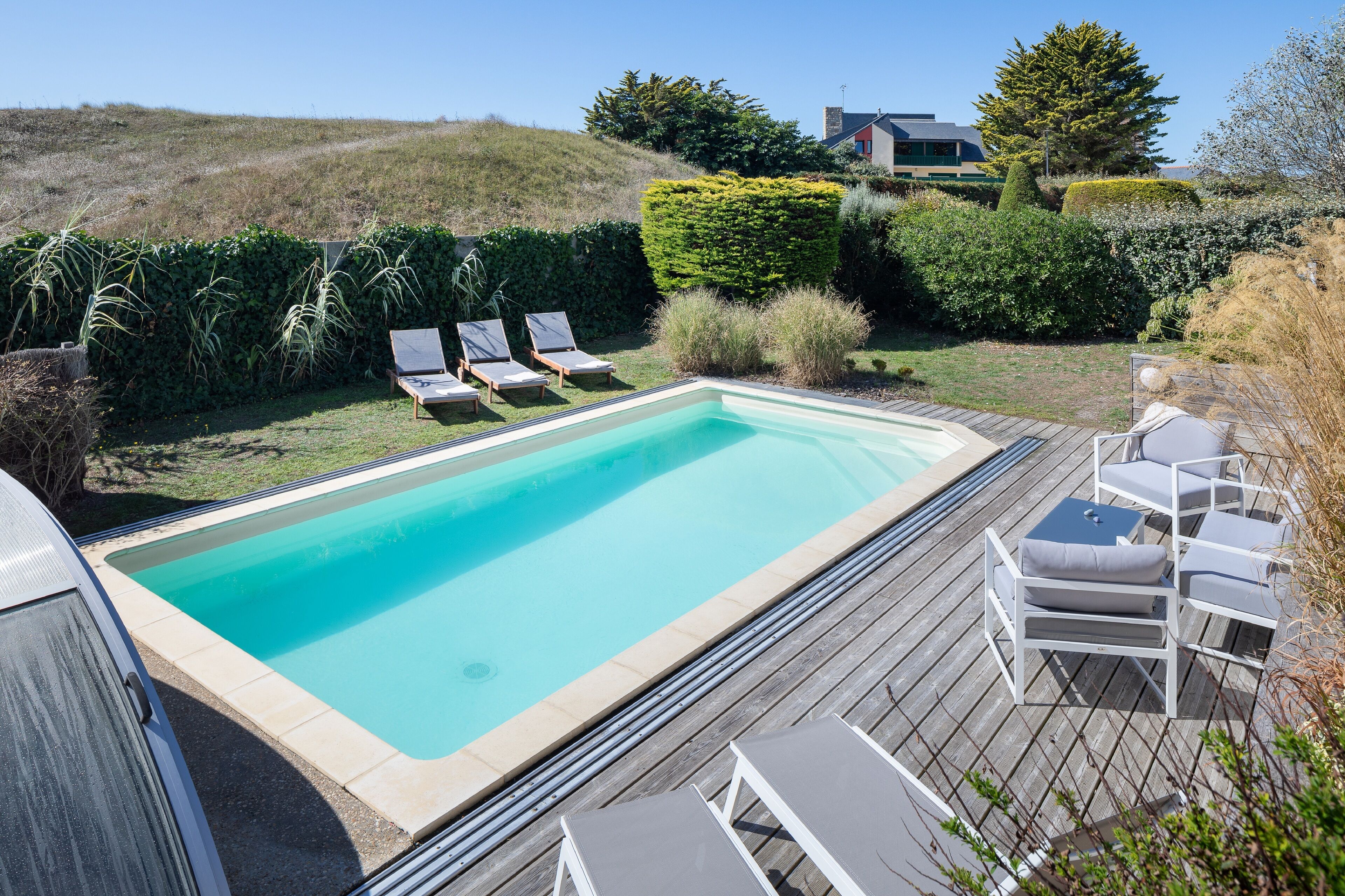 Ein rechteckiger Pool mit Holzterrasse, umgeben von Liegestühlen und einer Hecke.