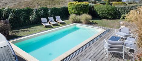 Ein rechteckiger Pool mit Holzterrasse, umgeben von Liegestühlen und einer Hecke.
