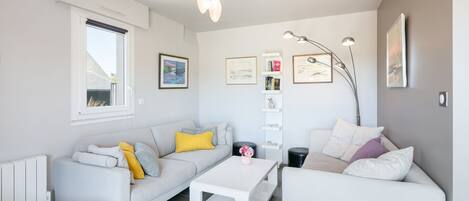 A modern living room with a white sofa, a coffee table, and a floor lamp. There are framed pictures on the wall and a radiator on the left.