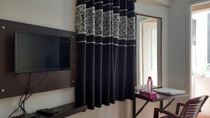 Une chambre dotée d’un téléviseur à écran plat fixé au mur, d’une petite table avec une chaise et d’une fenêtre avec des rideaux noirs.