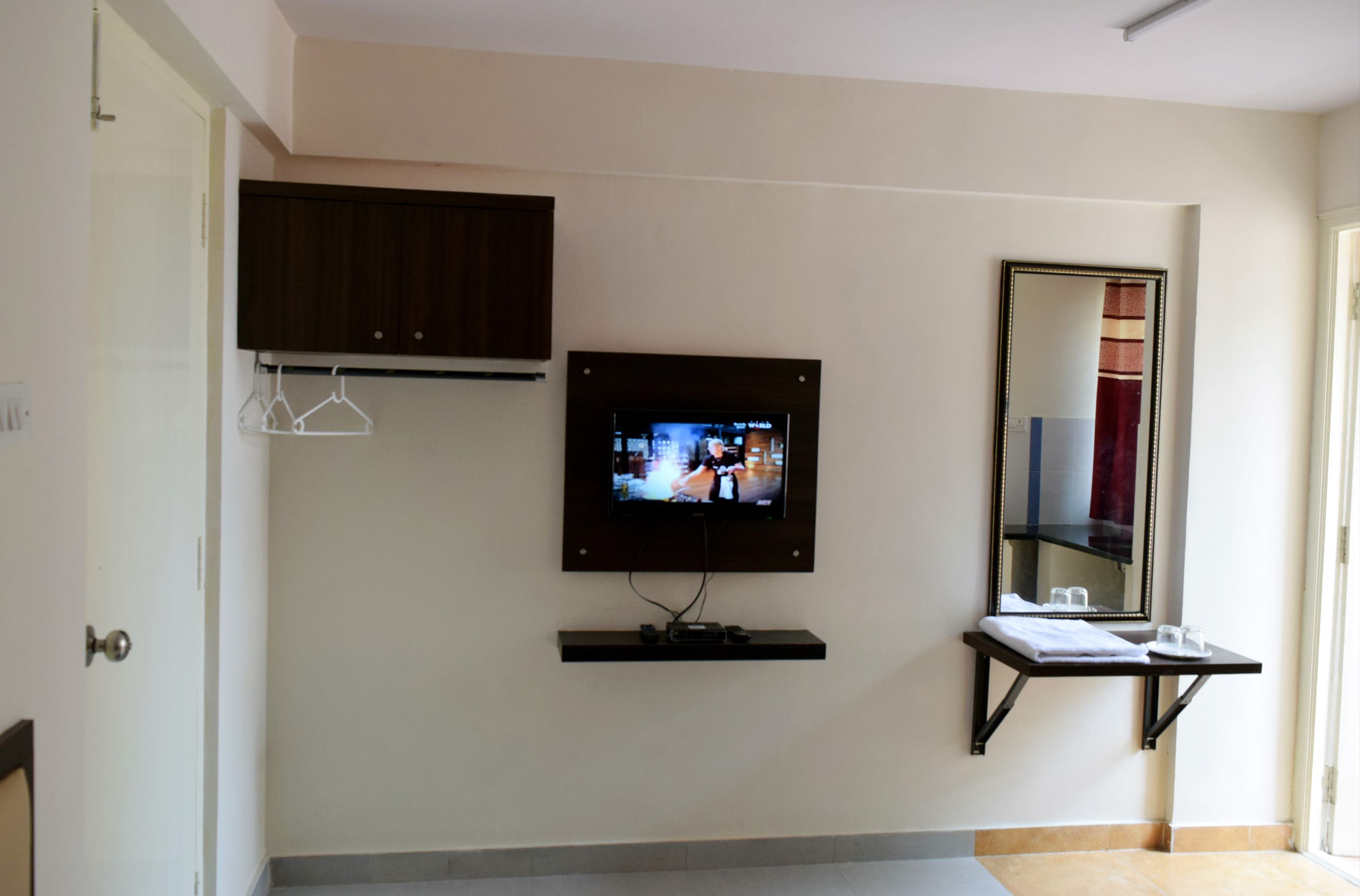 A hotel room with a television mounted on the wall, a mirror above a small table, and a door to the left.
