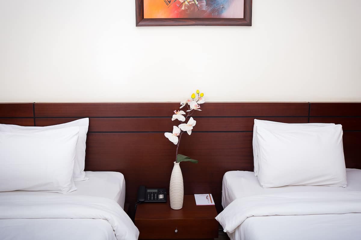 A hotel room with two beds, a wooden headboard, a white vase with flowers, a bedside phone, and a framed picture on the wall.