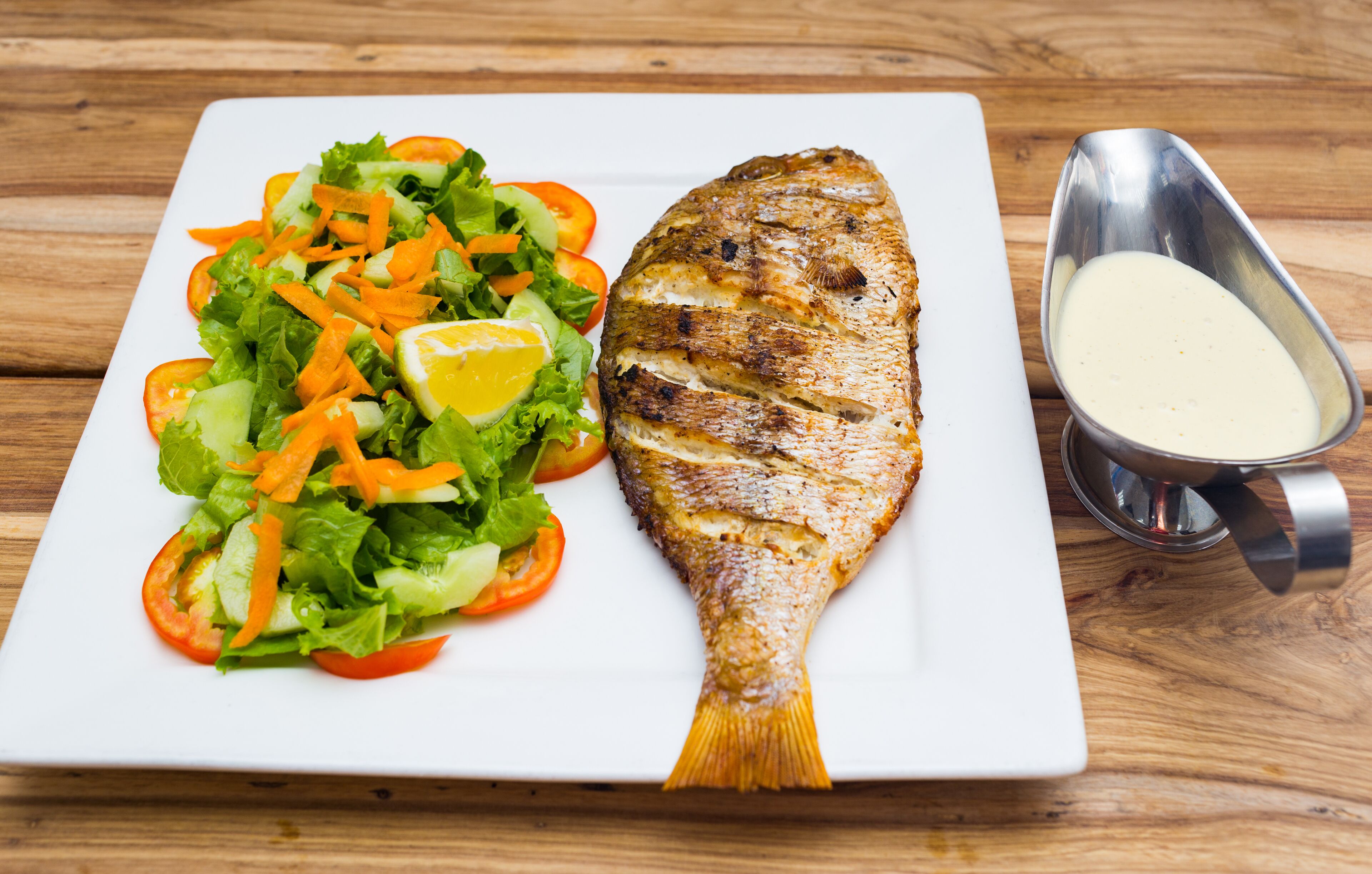 Une assiette blanche contenant du poisson grillé, une salade et un petit bol de sauce, le tout posé sur une table en bois.