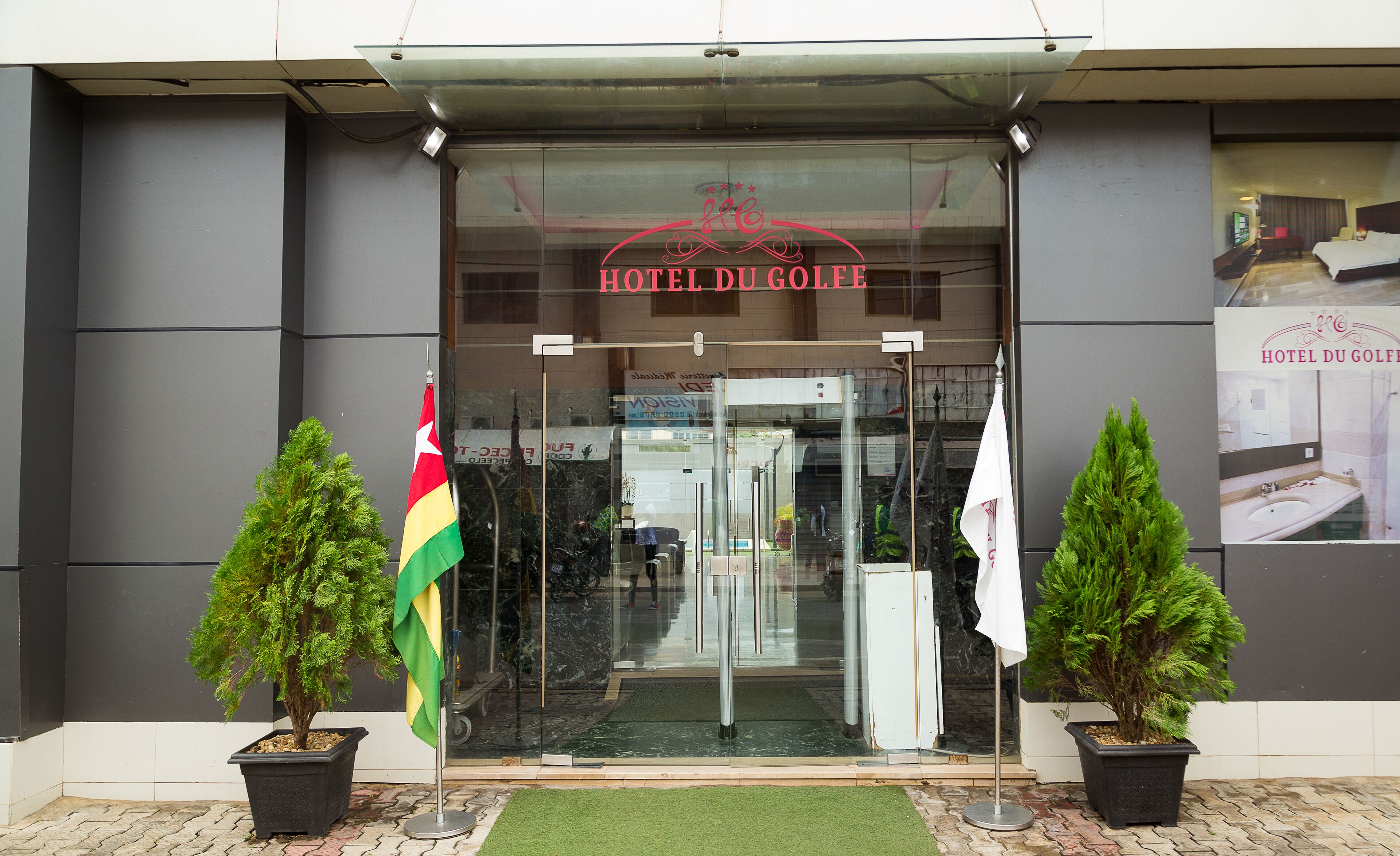 The entrance to Hotel Du Golf with a glass door, potted plants, and flags.