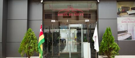 The entrance to Hotel Du Golf with a glass door, potted plants, and flags.