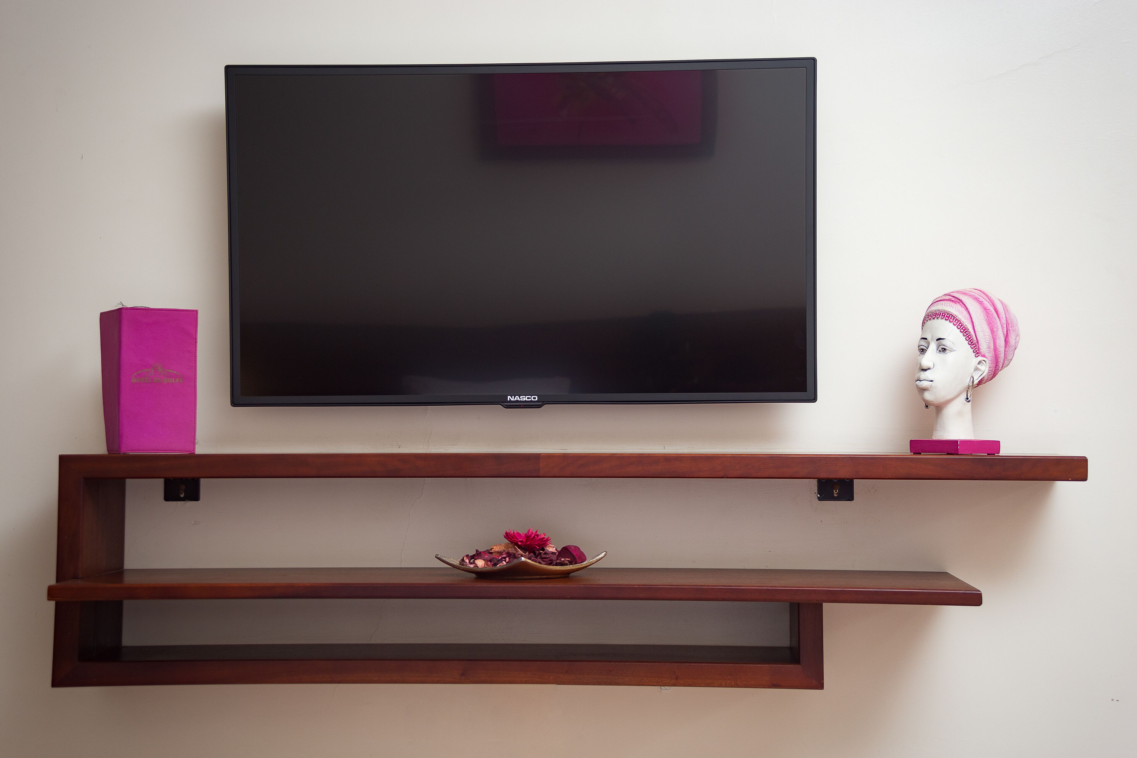A flat-screen TV mounted on a wall above a wooden shelf with a pink box and a bust sculpture.