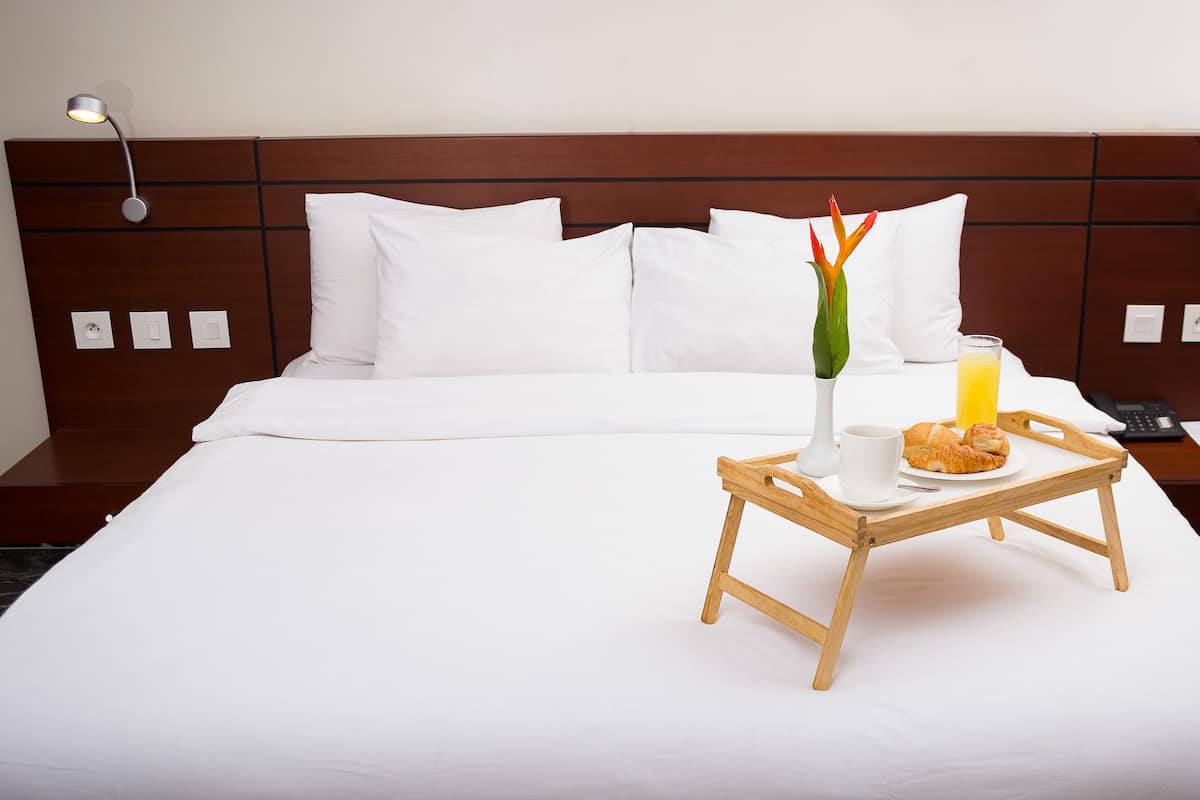 A neatly made bed with white linens, a bedside table with a lamp, a tray with croissants, a glass of orange juice, and a vase with flowers.