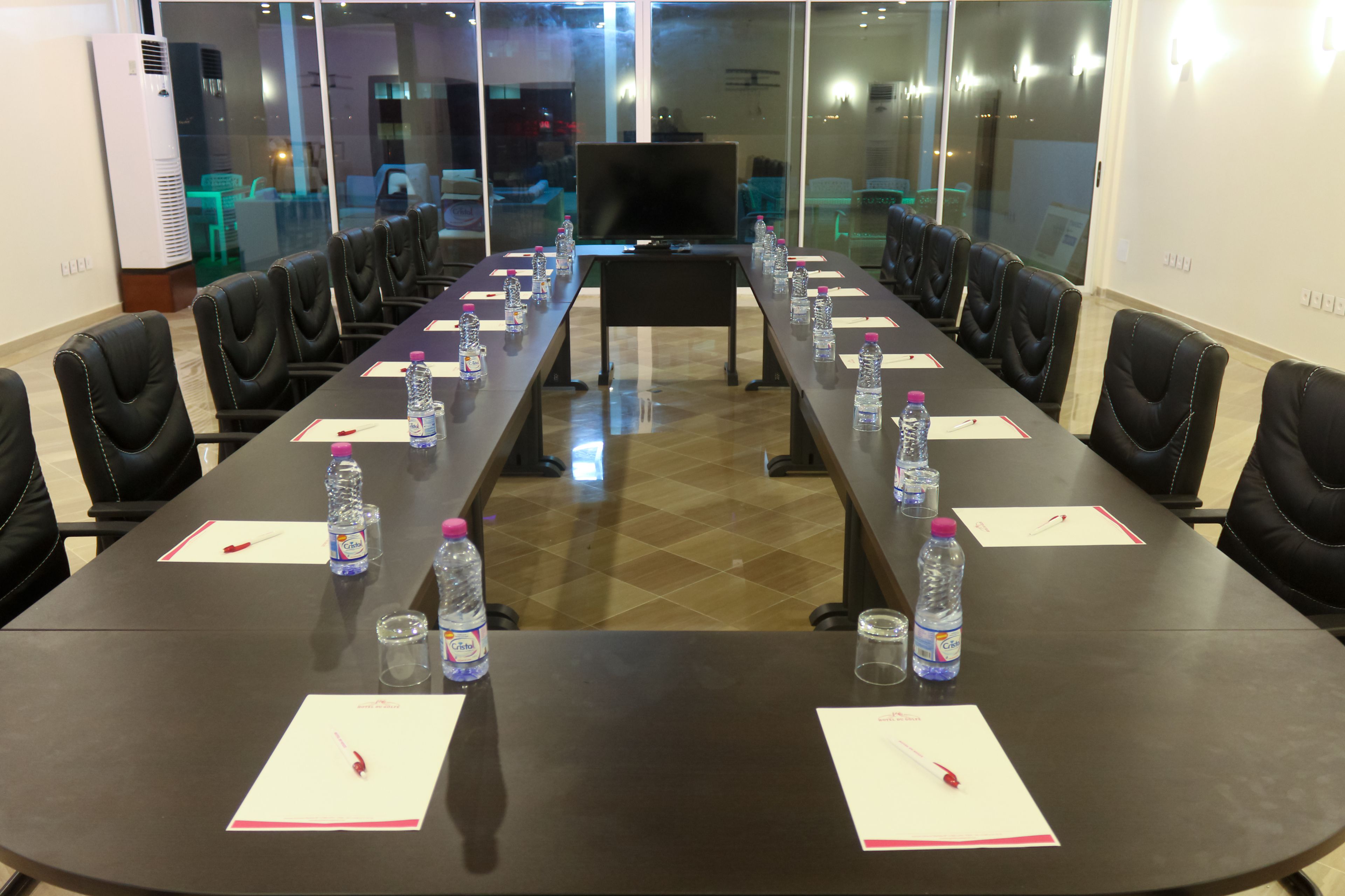 Un salón de conferencias con una mesa larga, sillas, botellas de agua y una televisión.