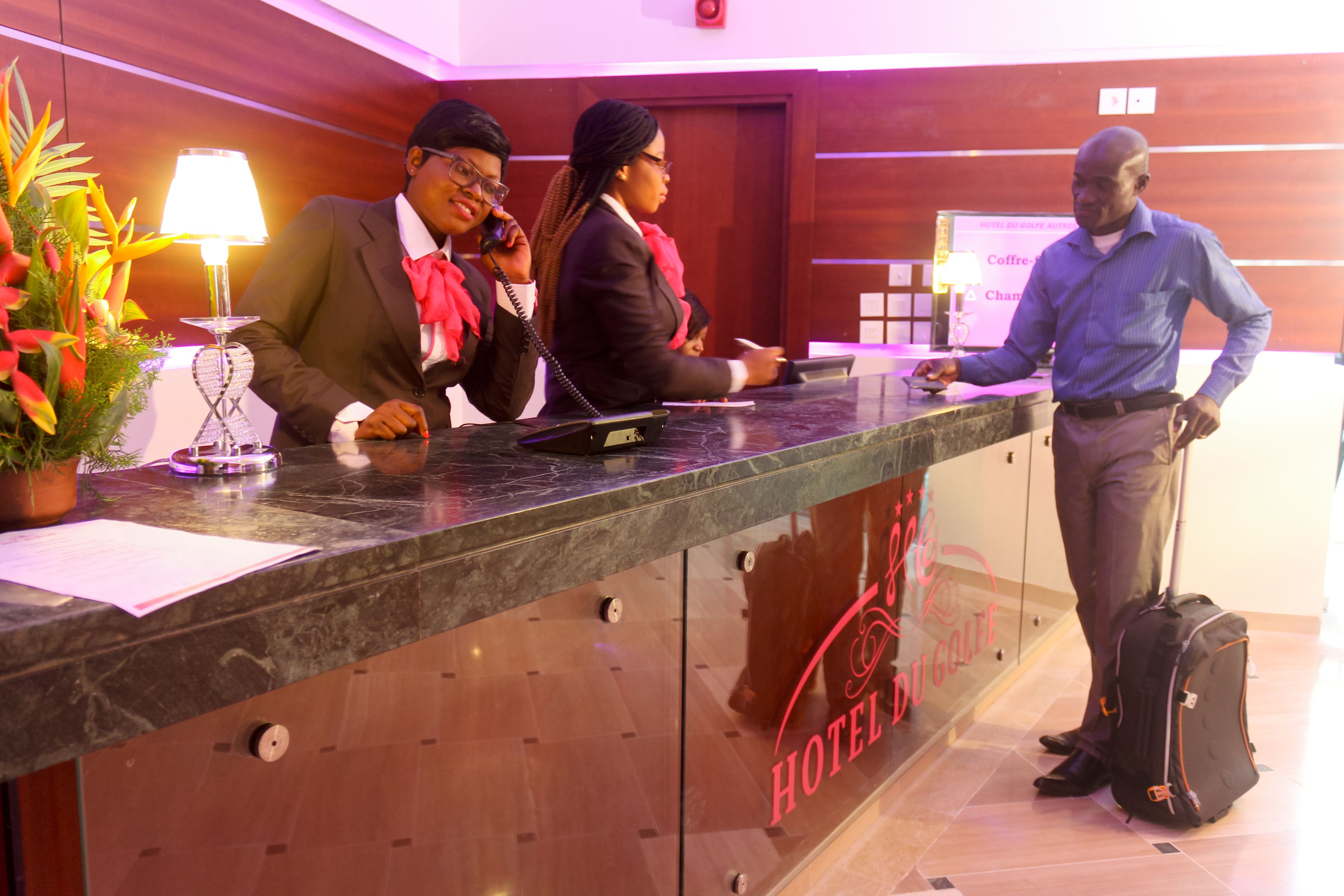 A hotel reception desk with a glass partition, a red logo, and a potted plant.