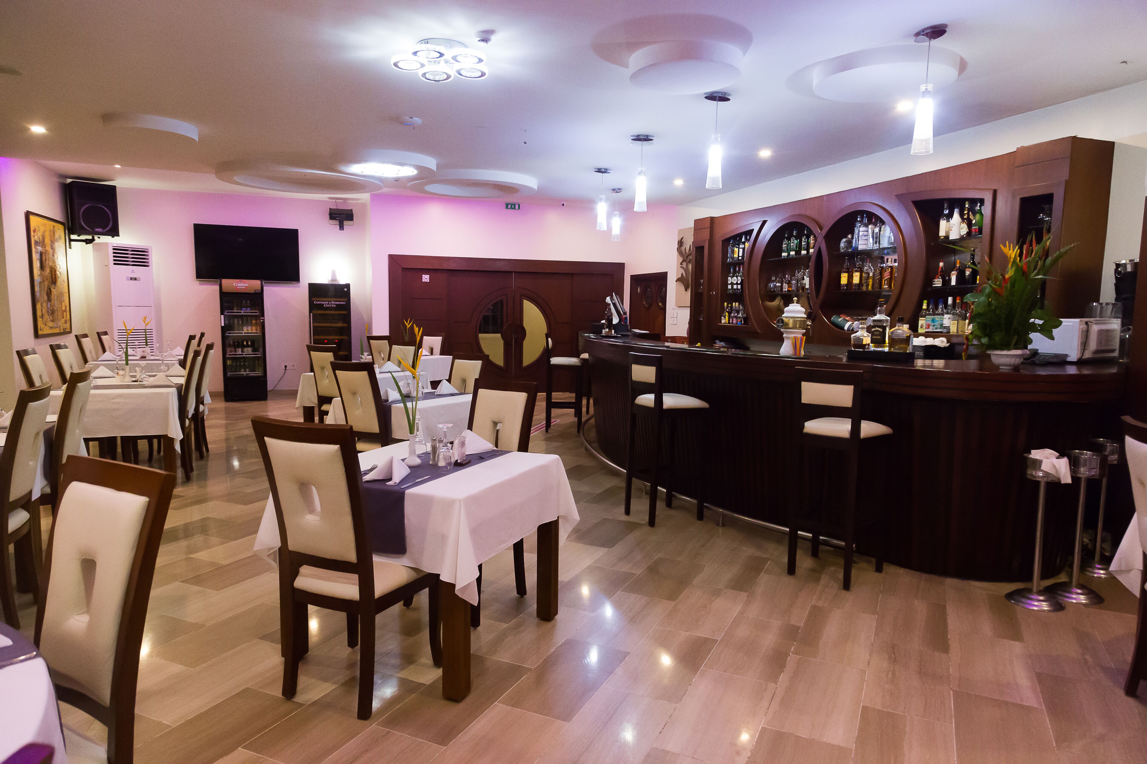 A restaurant with multiple tables and chairs, a bar area with shelves of bottles, and a television mounted on the wall.