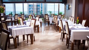 A restaurant with multiple tables and chairs, large windows with a view of the sea, and a speaker mounted on the wall. - Hôtel du Golfe (Lomé)