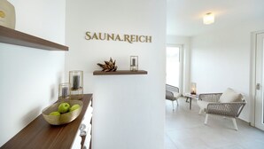 A modern interior with a sign reading "SAUNA.REICH" on the wall, a wicker chair, a small table with a candle, and a wooden shelf with decorative items. - Kucher's Genuss & Businesshotel (Darscheid)