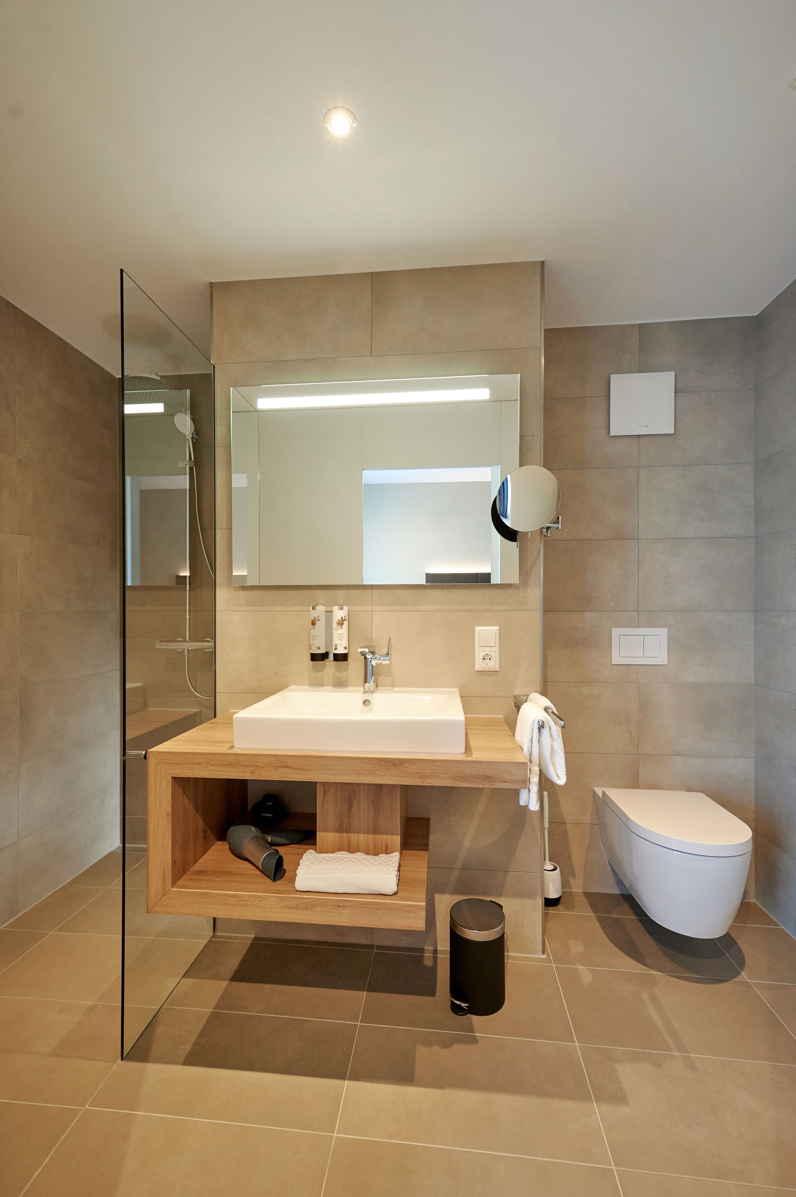 A modern bathroom with a glass shower enclosure, a wooden vanity, a white sink, a toilet, and a large mirror.