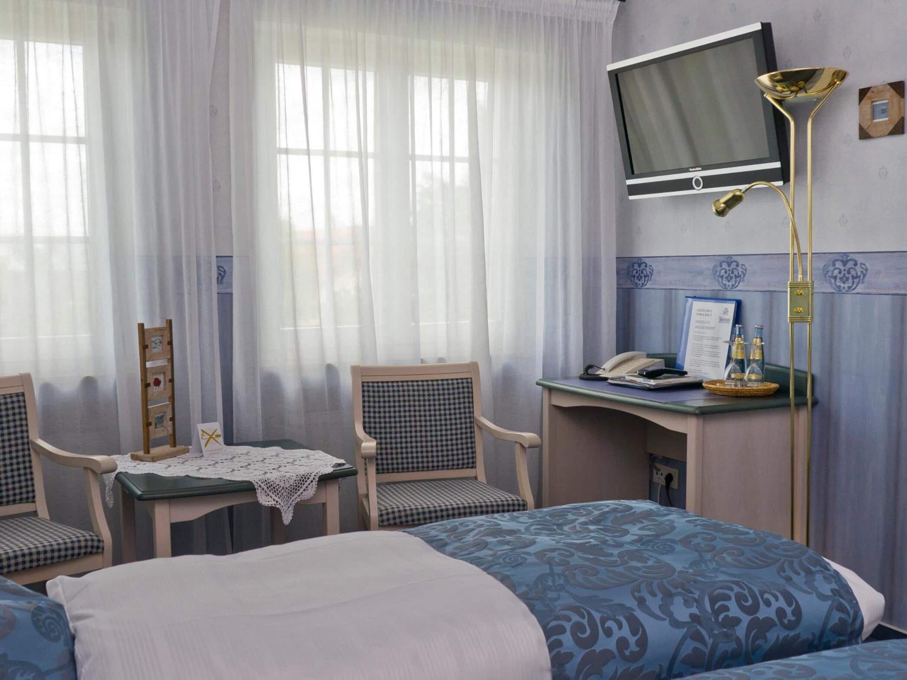 A hotel room with two beds, a desk with a telephone and a framed certificate, a chair, a small table with a lace tablecloth, and a television mounted on the wall.
