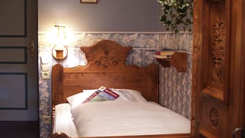 Ein Bett mit Holzbettgestell, ein Nachttisch mit Lampe sowie eine Wand mit Blumenmuster-Tapete und gerahmten Bildern.