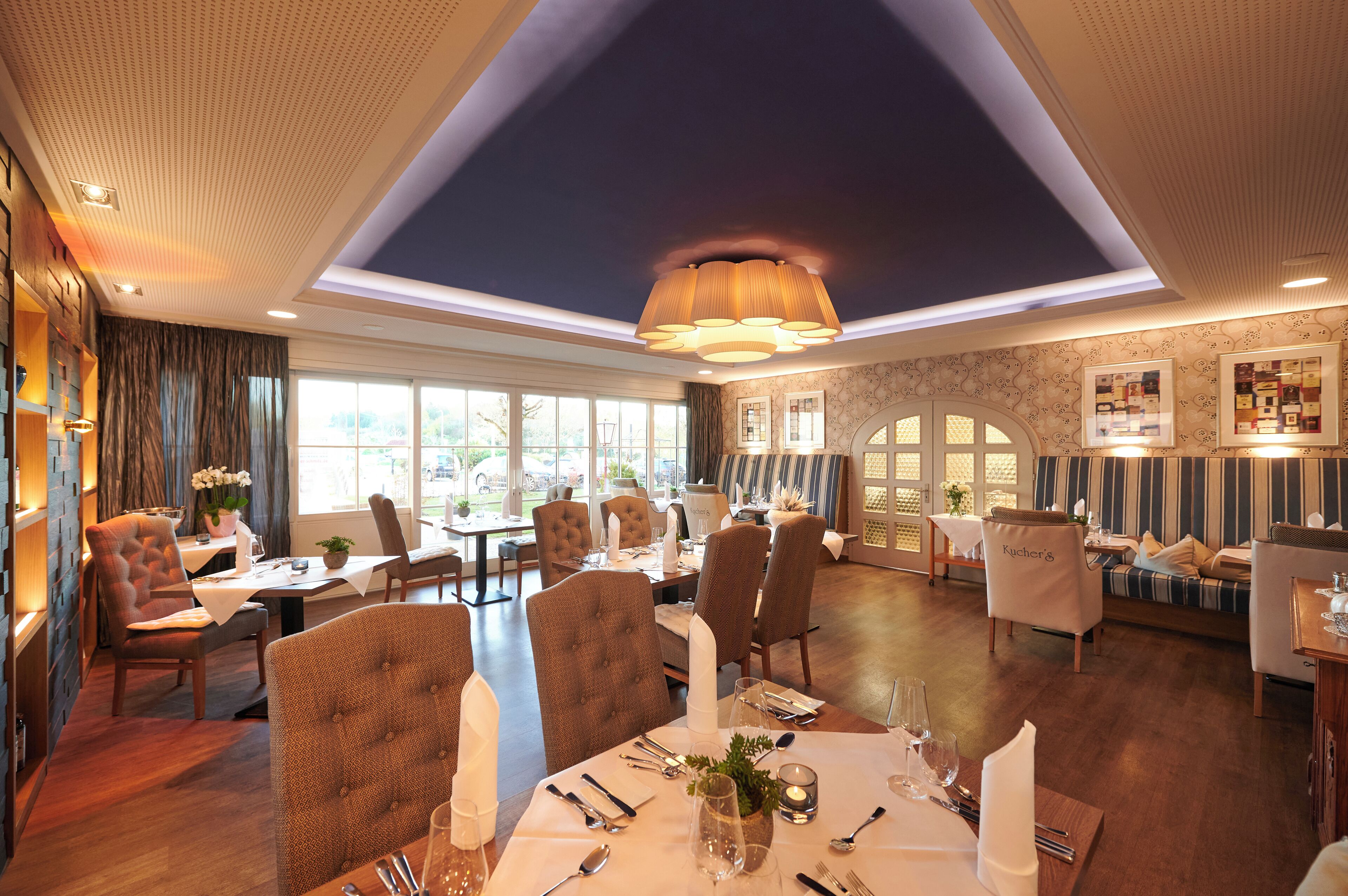 A dining area with tables set for four, upholstered chairs, and a large chandelier.