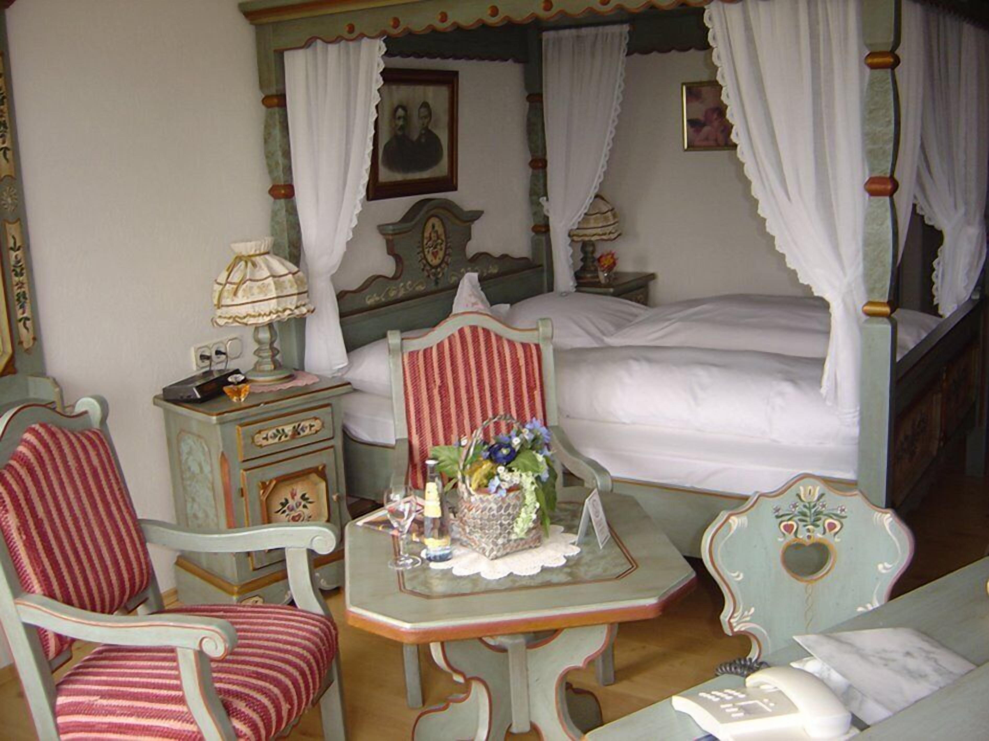A bedroom with a bed, two chairs, a nightstand, a table with a lamp, and a picture frame on the wall.
