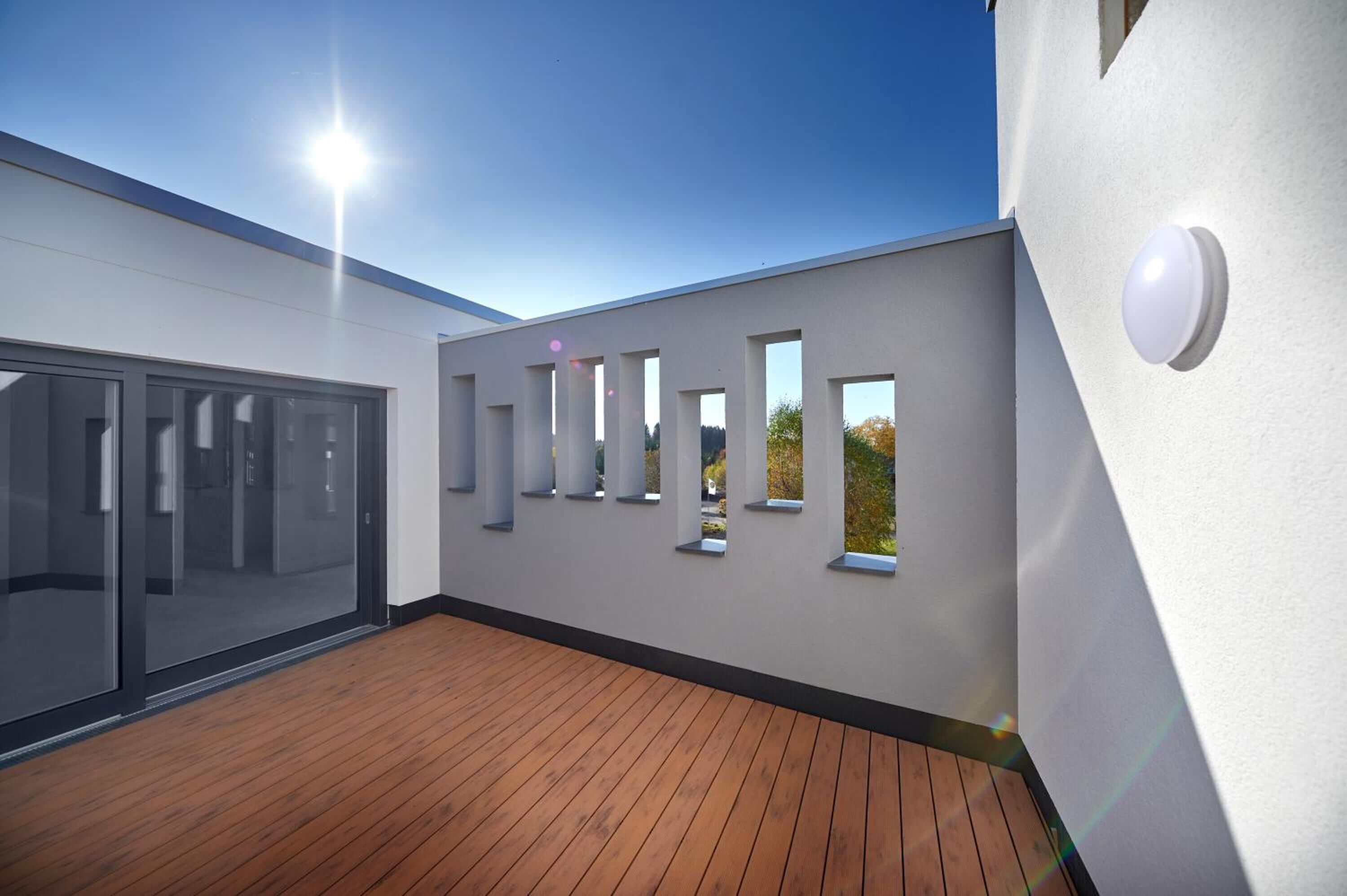 Fachada moderna de un edificio con terraza de madera, ventanas amplias y cielo despejado.