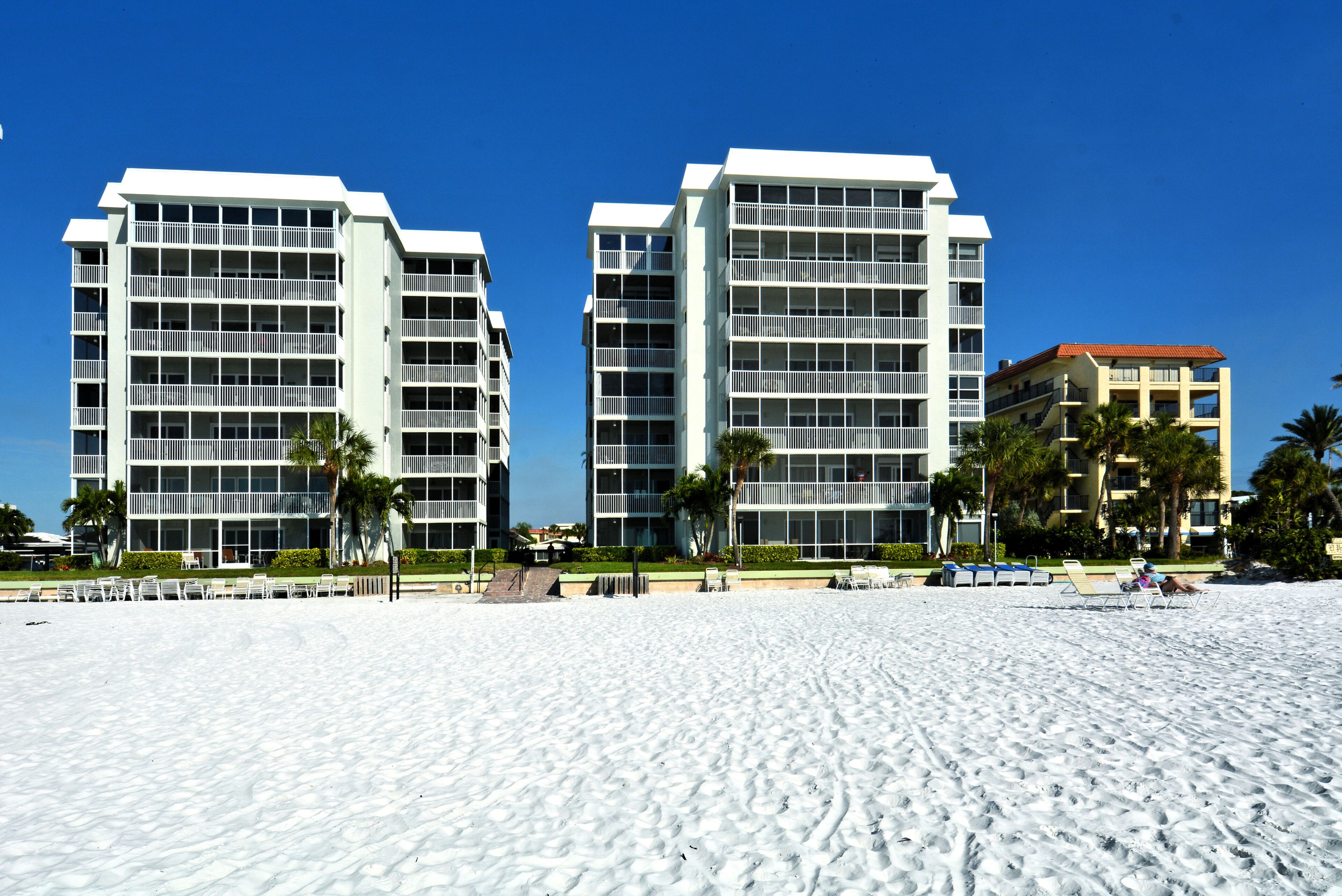 A beachfront hotel with multiple floors, balconies, and a sandy beach area with sun loungers.