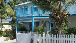 Un bâtiment à deux étages, avec une clôture blanche en bois, un balcon doté de balustrades bleues et un palmier au premier plan.