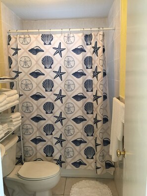 Une salle de bain avec un rideau de douche orné d’un motif représentant des coquillages et des étoiles de mer, des toilettes, un porte-serviettes avec des serviettes pliées, et un tapis blanc sur le sol.