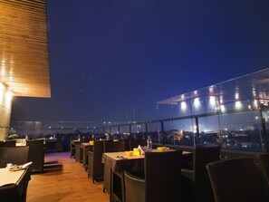 Un espacio de comedor al aire libre con mesas y sillas, vista a la ciudad y un amplio ventanal.