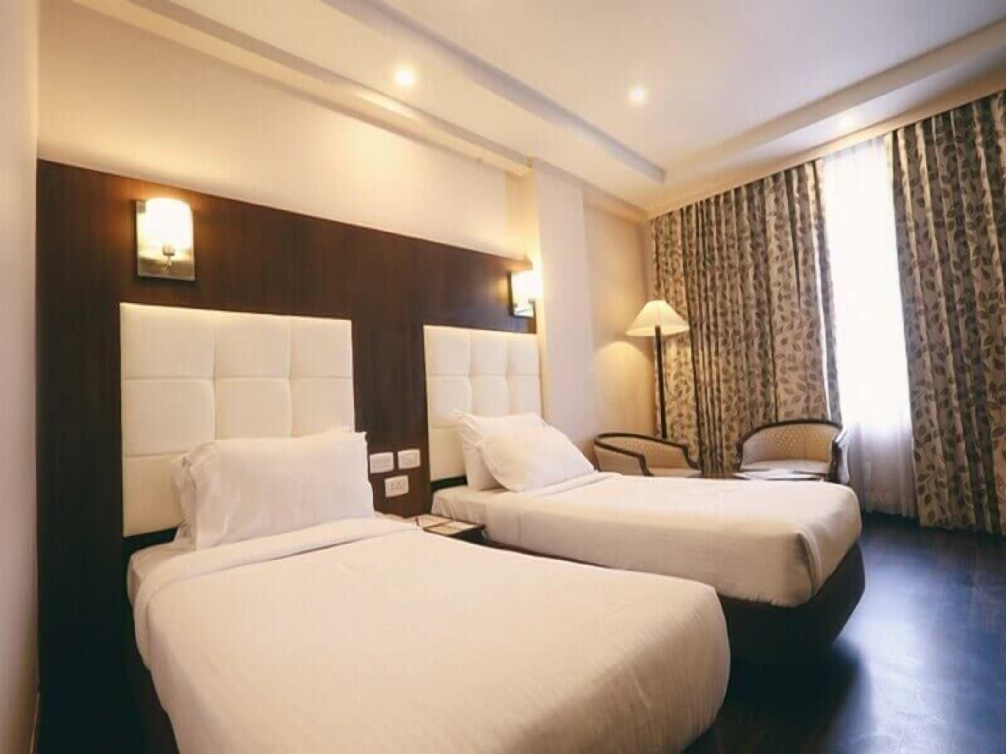 Habitación de hotel con dos camas, una mesita de noche, una lámpara y una ventana con cortinas.