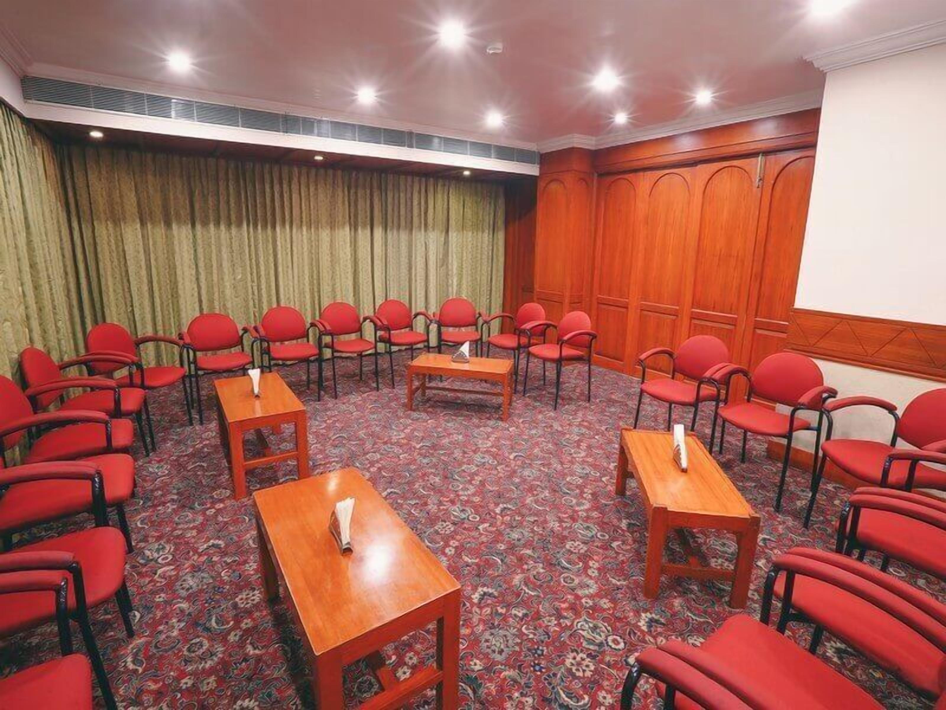 A conference room with a carpeted floor, red chairs, wooden tables, and a projector screen.