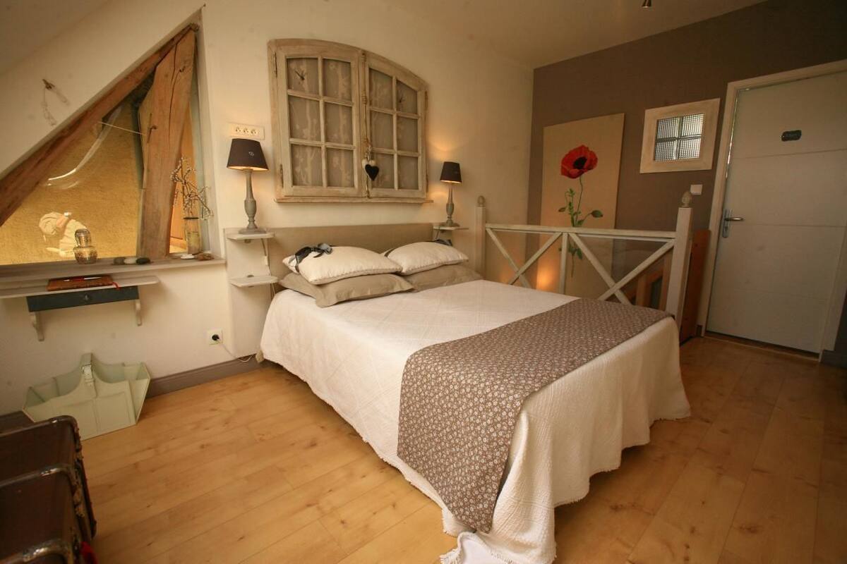 A bedroom with a bed, a window, a lamp, a door, and a wall with a flower painting.