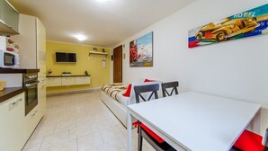 Een compacte keuken met een magnetron, oven en spoelbak. Er is een eethoek met een witte tafel en rode stoelen. Op de muur hangt een schilderij van een auto.