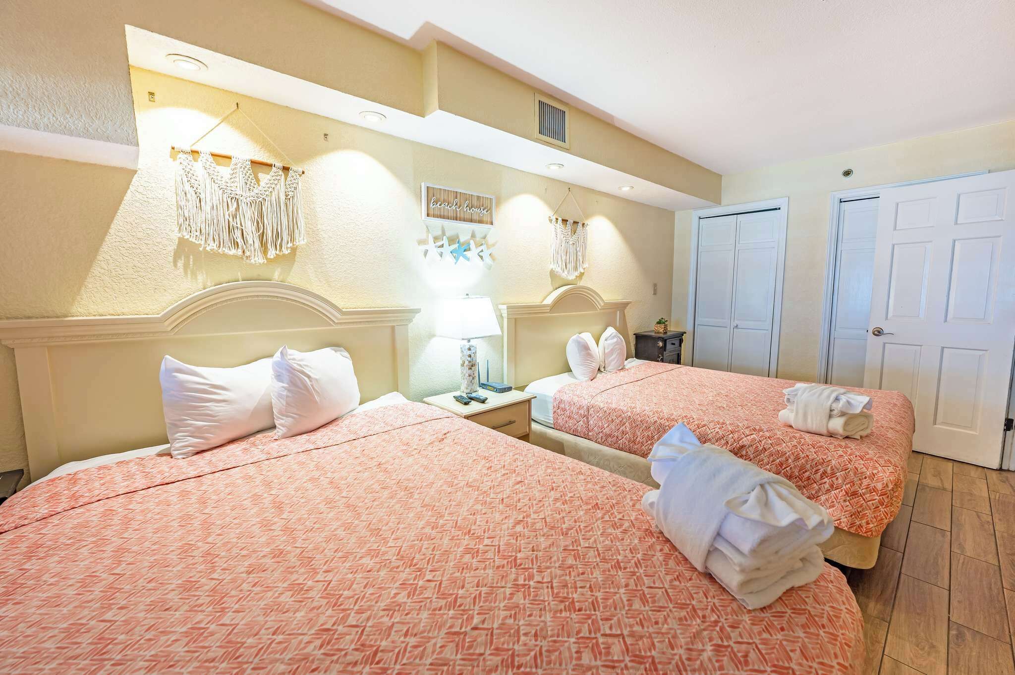 Une chambre d’hôtel avec deux lits, une table de chevet avec une lampe, un miroir et une décoration murale en macramé.